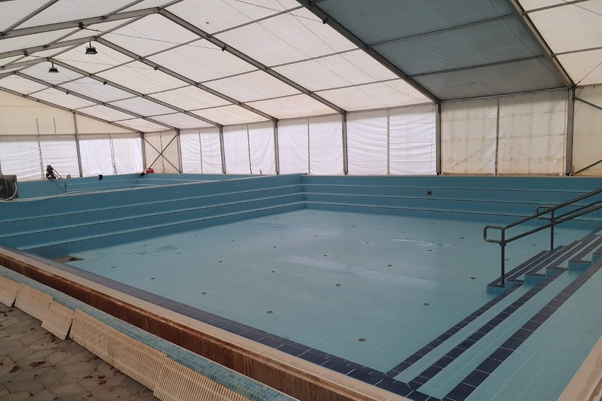 Felújítás alatt álló nagyméretű uszodai medence világoskék csempeburkolattal, sátorral fedett munkaterületen.